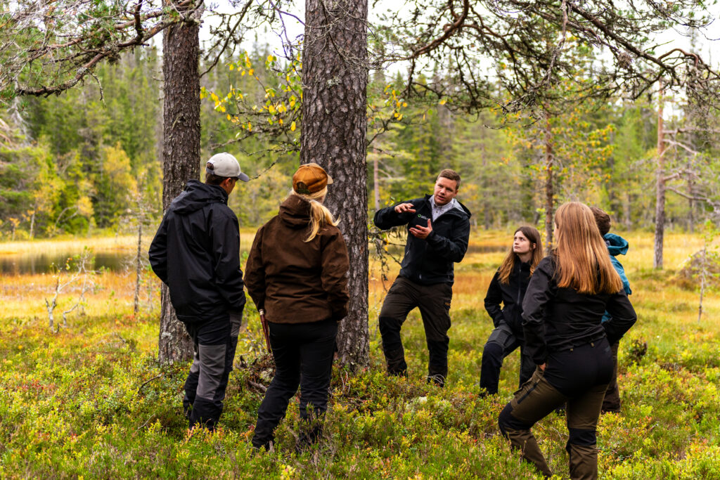 Lektion ute i skogen, på Älvdalens Utbildningscentrum. En lärare står vid en gammeltall och berättar, elever lyssnar.